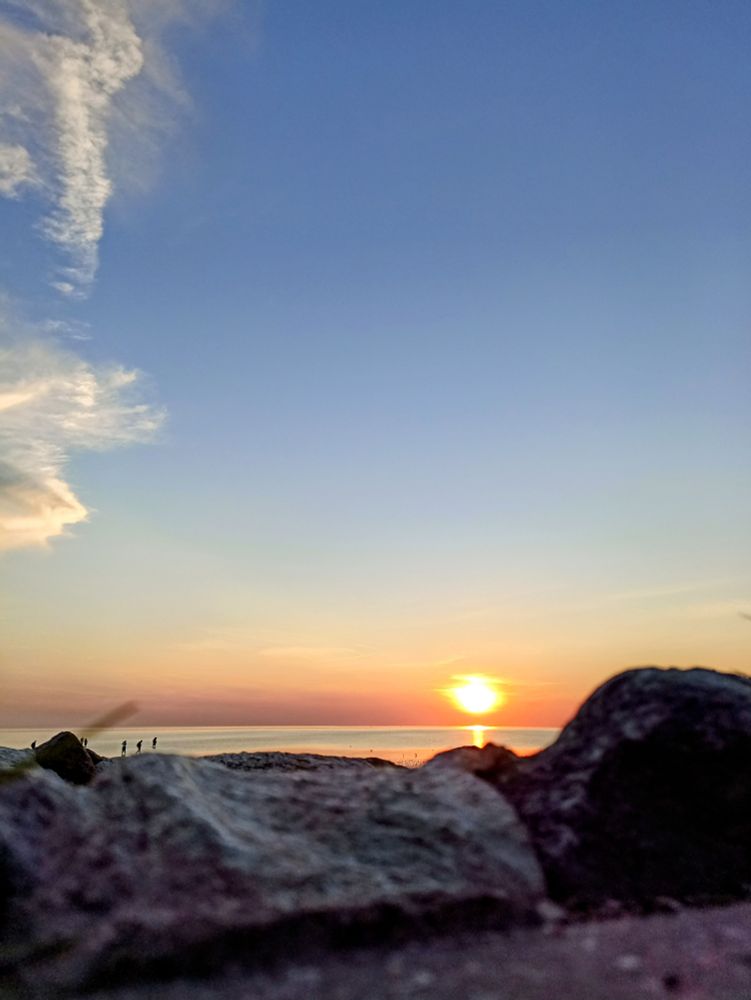 im Vordergrund Steine, dahinter Meer, untergehende Sonne, Bauer Himmel 