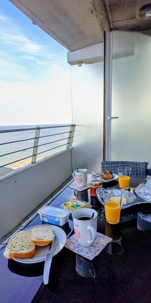 ein Tisch mit Tellern, Tassen, Gläsern, Brötchen, Aufstriche; Blick über ein Balkongeländer auf das Meer 