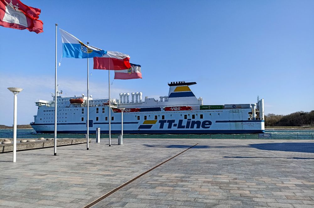 eine Fähre mit der Aufschrift TT-Line, im Vordergrund eine gepflasterte Fläche, links Fahnen, blauer Himmel 