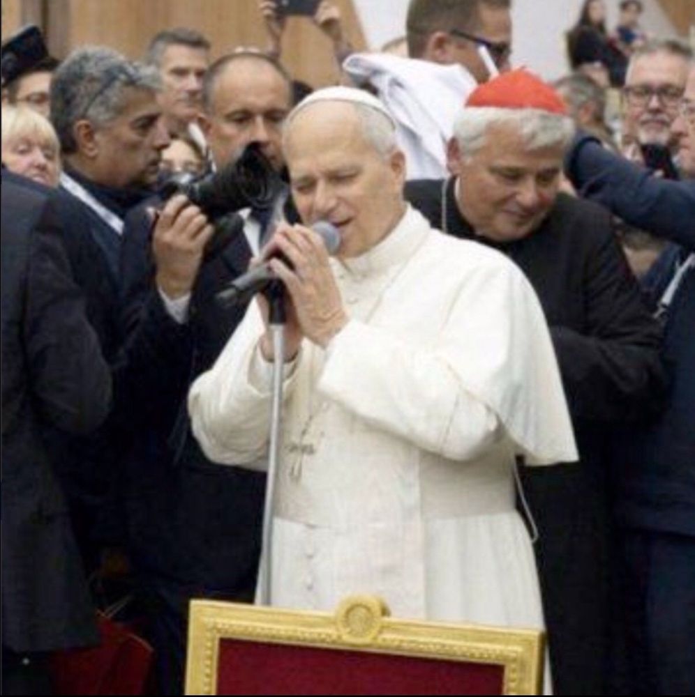 The pope dressed in white with his eyes closed and mouth open saying something into a microphone. Behind him are priests dressed in black