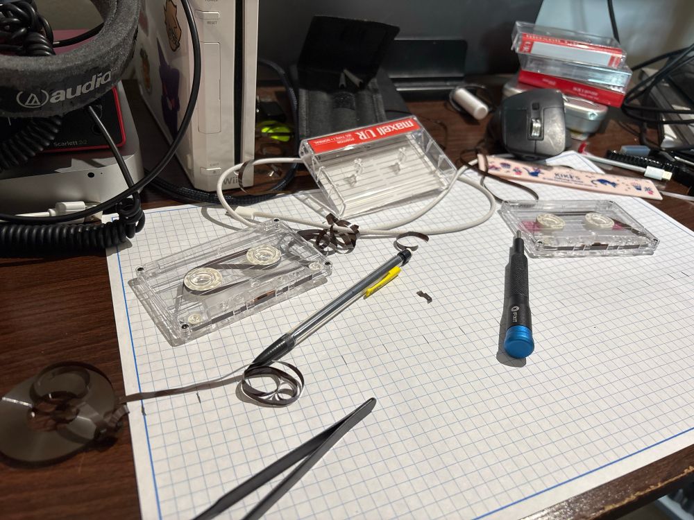 A photo of a cluttered desktop with multiple cassette tapes, tools and other electronics scattered about.