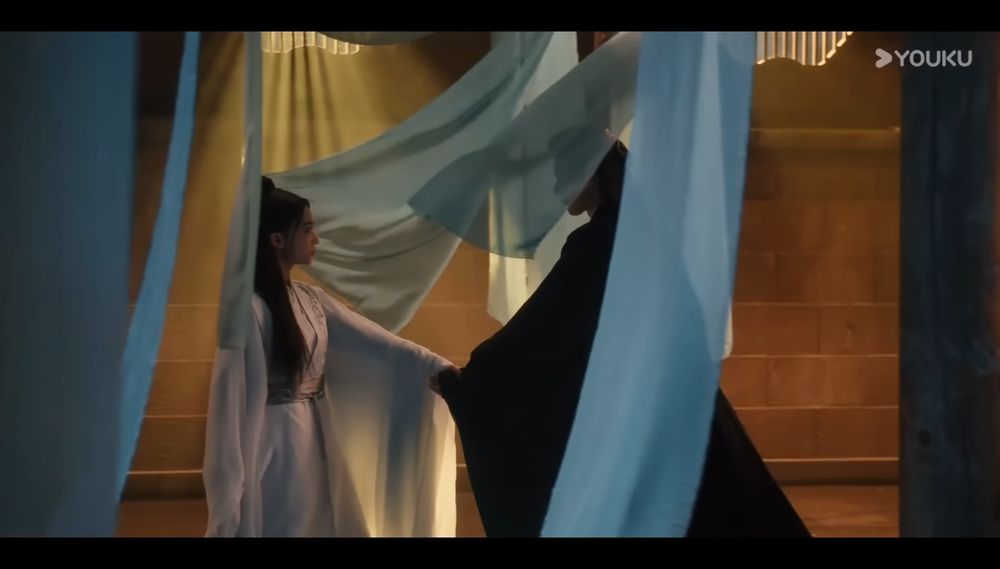 A still from the Chinese drama #KillMeLoveMe showing a man and a woman holding hands. The woman is leading the man through light blue & green hanging fabrics. 