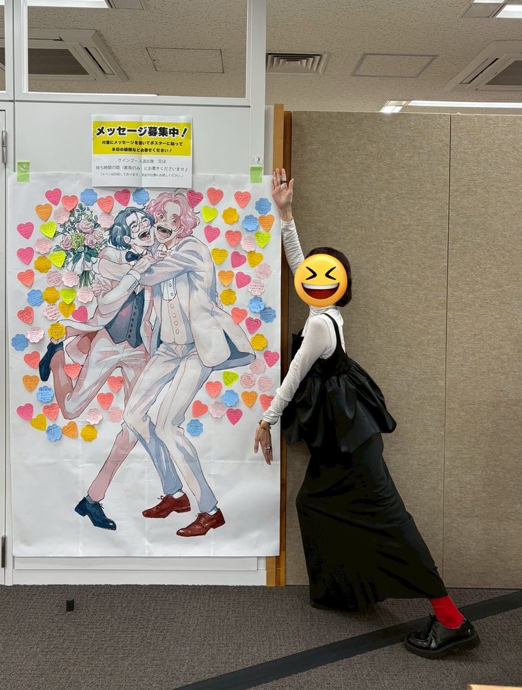 A photo of Mone Sorai with her characters from the manga Our Not-So-Lonely Planet Travel Guide. She is wearing a white turtleneck, black ruffled sleeveless shirt over top, long black skirt, red socks and black shiny Oxford shoes. 