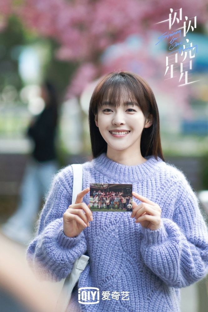An image from the Chinese drama #ShiningForOneThing showing a young woman wearing a light purple sweater holding a photo of a group of children. She is smiling. 