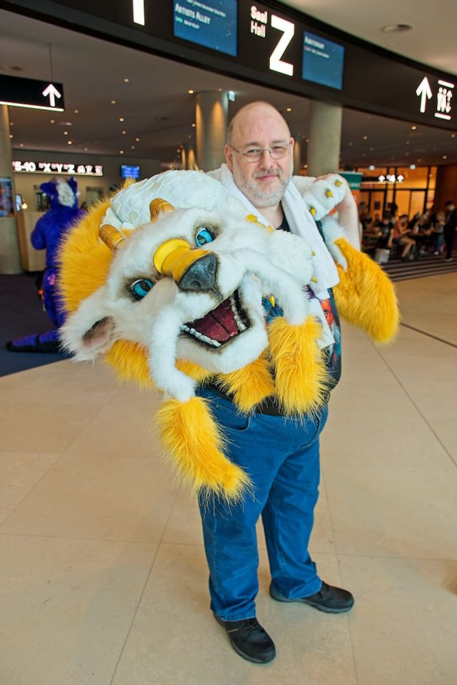 Taken in the Entrace Hall of the CCH during Eurofurence 2026. Whitepelt holding Kingane, a Dragon Puppet with white fur, yellow golden Tufts and blue eyes looking directly into the camera