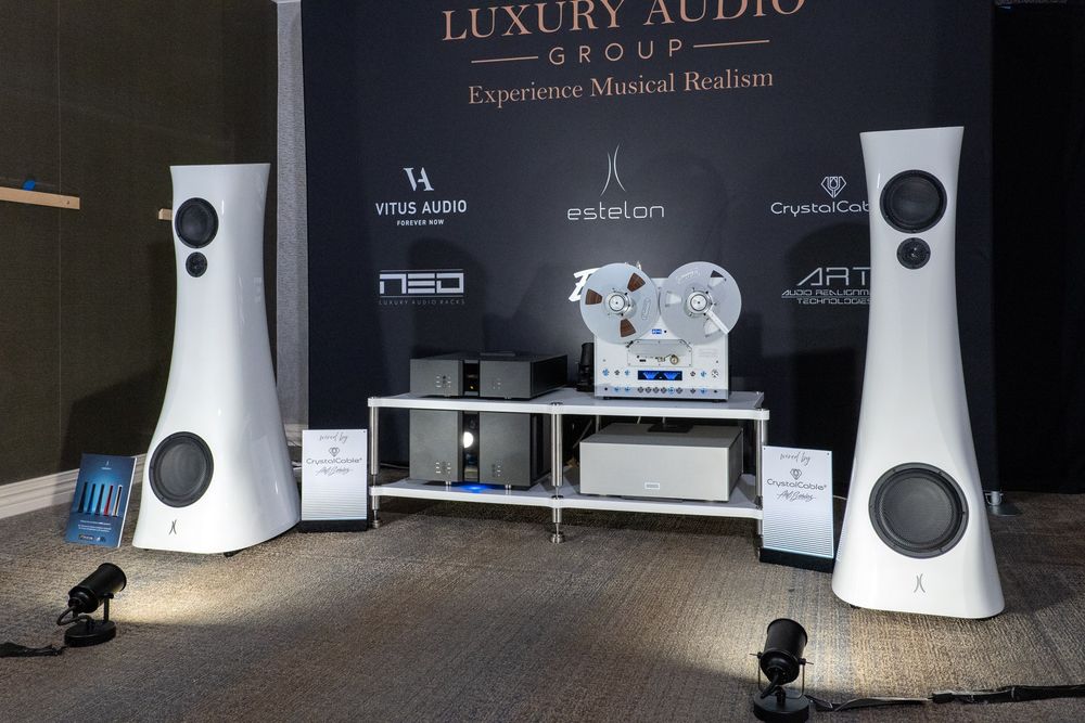The image showcases a high-end audio system displayed by the Luxury Audio Group, with a backdrop reading "Experience Musical Realism." The centerpiece features a sleek reel-to-reel tape deck positioned on a minimalist rack, flanked by black and silver audio components, including amplifiers and power supplies. On either side of the setup stand two Estelon loudspeakers, distinguished by their curved, glossy white enclosures with integrated drivers, designed for premium sound performance. Spotlights enhance the aesthetic appeal of the speakers and equipment. Logos for brands such as Vitus Audio, Estelon, NEO, ART, and CrystalCable are visible in the background, emphasizing the luxury and exclusivity of the system.