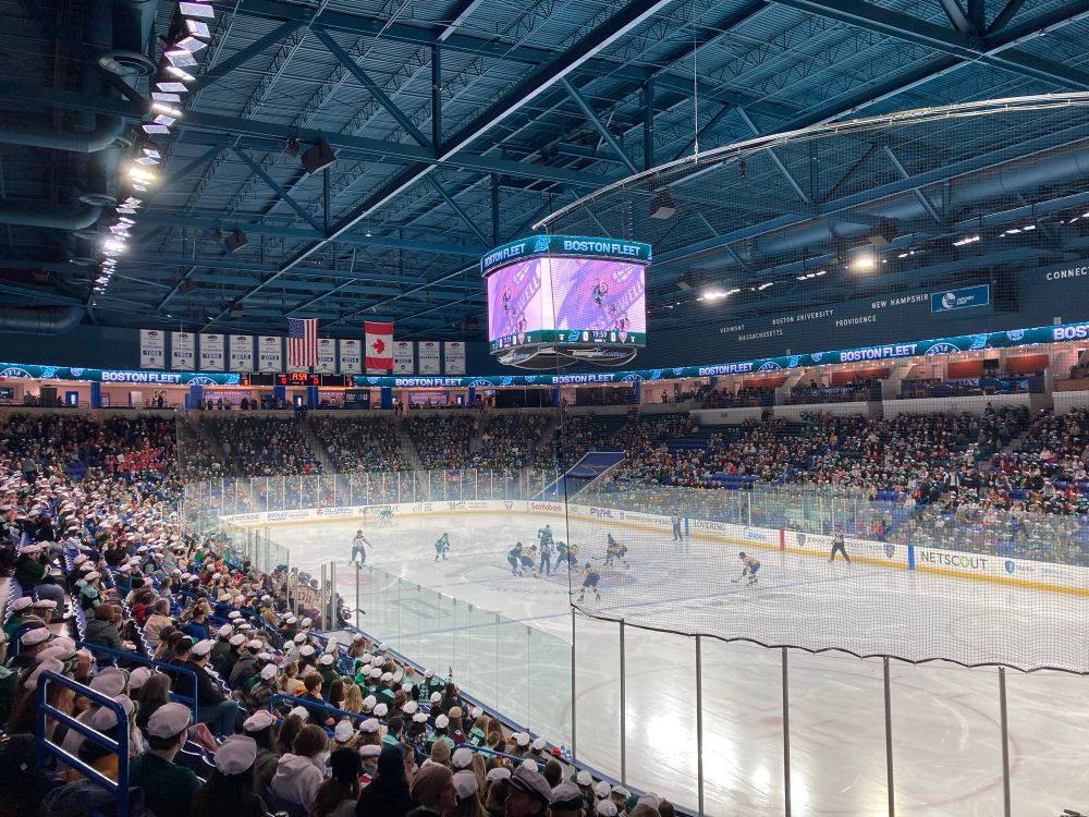 View of an indoor hockey rink with players from two teams positioned on the ice. The stands are packed with fans. The Jumbotron display says “Boston Fleet”.