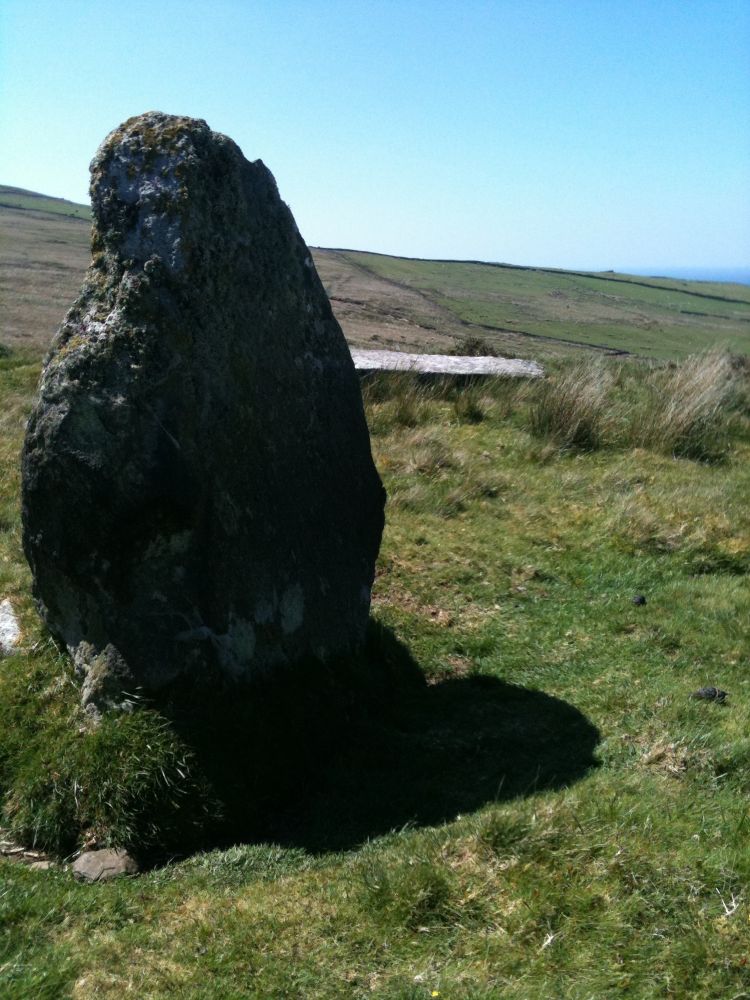 A dark moss- and lichen-covered squat troll of a stone casts a short black shadow over the scrubby moorland grass in the noon sunshine on a cloudless day. It appears as if it is  tipping its head quizzically, whilst behind it lies a long, pale grey massive toppled stone. There must be some connection here between these very different neighbours that has remained in place for perhaps thousands of years, but the narrative lost… perhaps yet to be rediscovered?  To the left, the bleak  hillside slopes away upwards to a bright blue sky in the distance and, to the right, it drops away to reveal a bright blue sea. A place of many memories.