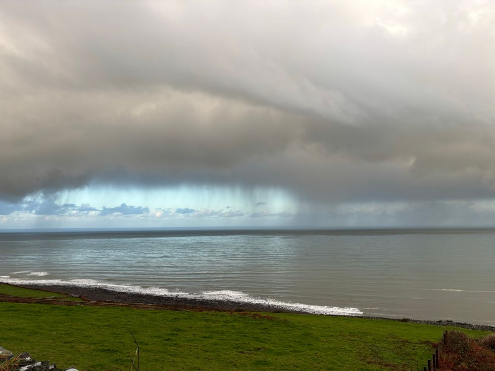 A wet green field slopes down to a grey stony beach with small white breaking waves on the shore.  The sea is calm and reflects the heavy dark grey clouds hanging low over the water. Wispy mares’ tails extend down from the clouds to brush a very thin dark grey cloud-shadow belt across the sea. Into the distance, beyond the heavy grey clouds and dark grey shadow on the sea, the sky and sea reflect brilliant blue back at each other. A claustrophobic sense of impending doom with escape visible but not reachable. 