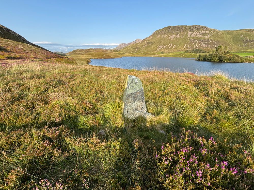 A hip height grey pointed standing stone stands on purple flowering moorland overlooking a deep blue lake with a small tree-covered island. Beyond, distant craggy mountains rise to a cloudless blue sky.