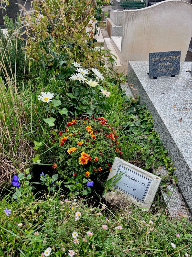 The grave of Jean Baudrillard in Montparnasse Cemetery in Paris. The grave is comprised of many bunches of flowers and vines and the memorial is inscribed on a small plaque placed within the flowers.