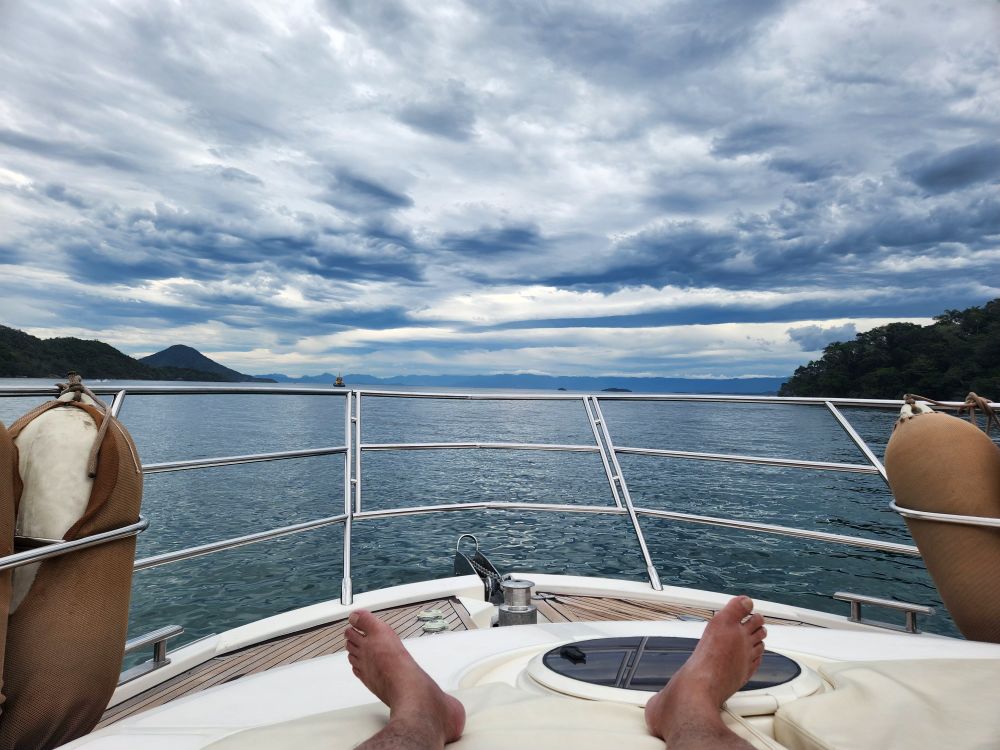 Foto tirada de uma das praias da Ilha Grande em direção ao continente. Na foto aparece meus pés, a ponta do iate, duas ilhas, o mar e o horizonte cheio de nuvens