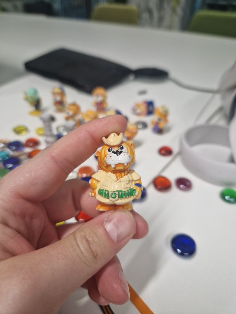 In my hand I hold a small plastic toy resembling a humanoid lion character. Cartoonish and dressed in a safari explorers garb. On the table in the background is spread a great number of colourful glass counters surrounding a few bits of blurry immersive technology.