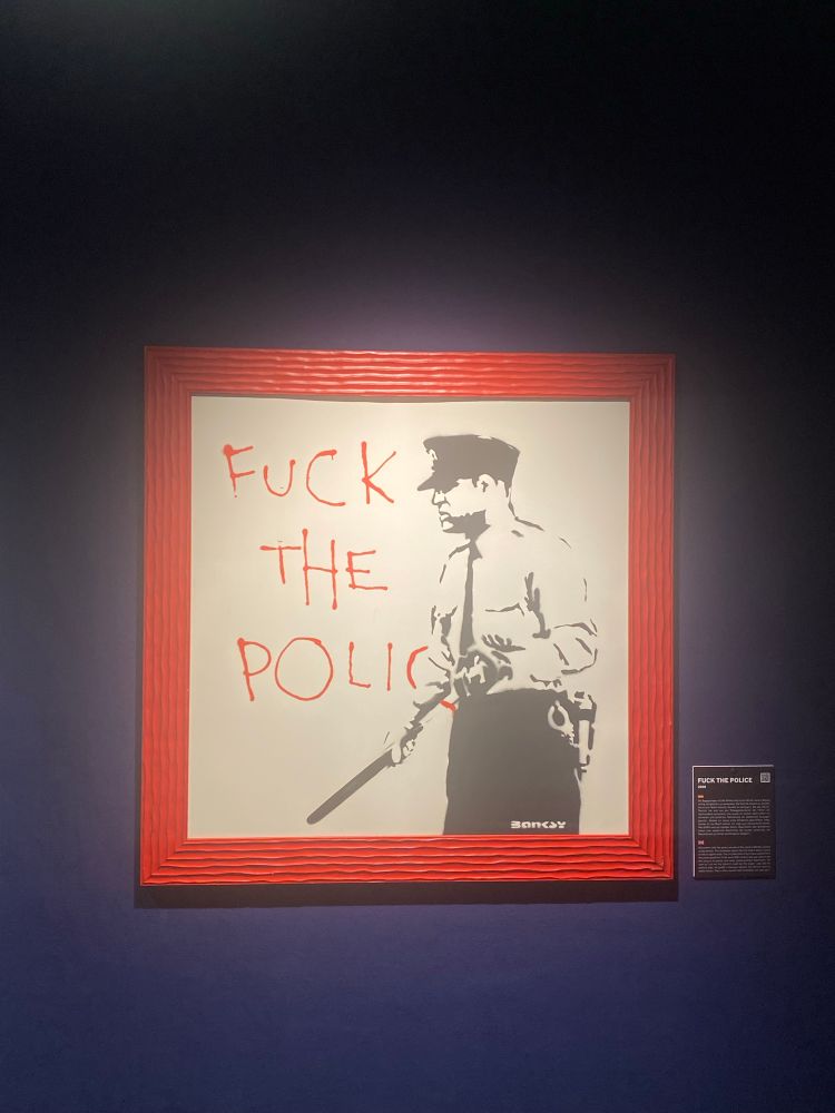 Ein in roten Rahmen gefasstes Bild von Banksy
Zu sehen ist ein schwarz-weiss Bild eines Polizisten mit gezücktem Schlagstock zu sehen, wo neben in roter Farben verfasster Tag "FUCK THE POLICE" steht