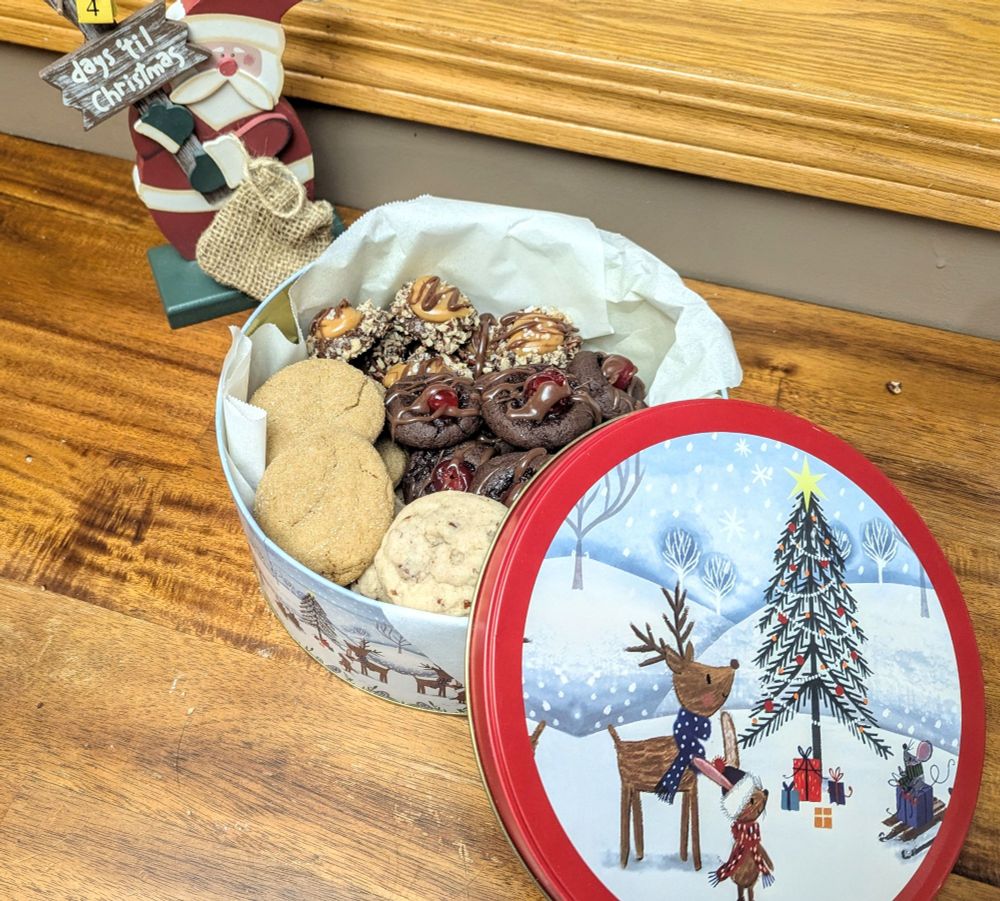 Photo of a Christmas themed cookie tin containing 4 kinds of cookies, ginger snaps, butter pecan, chocolate Cherry and turtle cookies. In the background is a "days 'til Christmas" wooden sign being held by Santa 