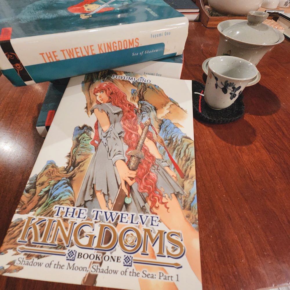 The new paperback edition of volume one of Fuyumi Ono's English translated novel series, The Twelve Kingdoms, laid against a stack of three original English versions of the same novel series