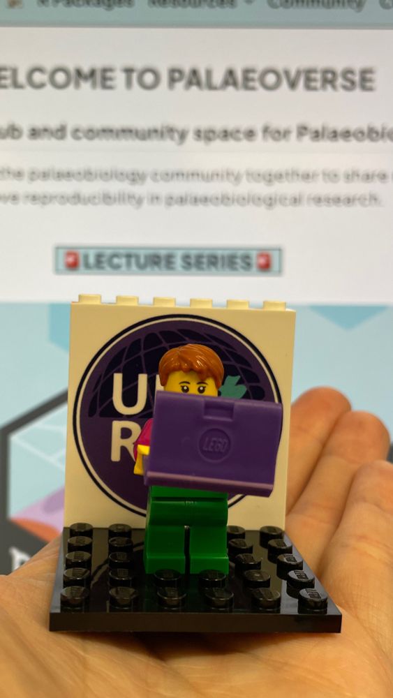 A lego minifigure of Dorothy Bishop with a laptop, which is a 'Doscar' award for the UKRN Dorothy Bishop Prize, held in an open hand, in front of a laptop screen with visible text "Welcome to the Paleoverse"