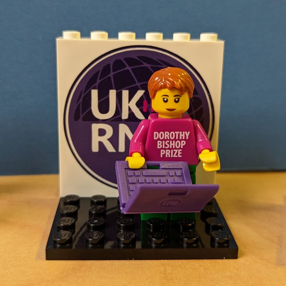 A lego minifig of Dorothy Bishop with a laptop, standing in front of the UK Reproducibility Network logo.