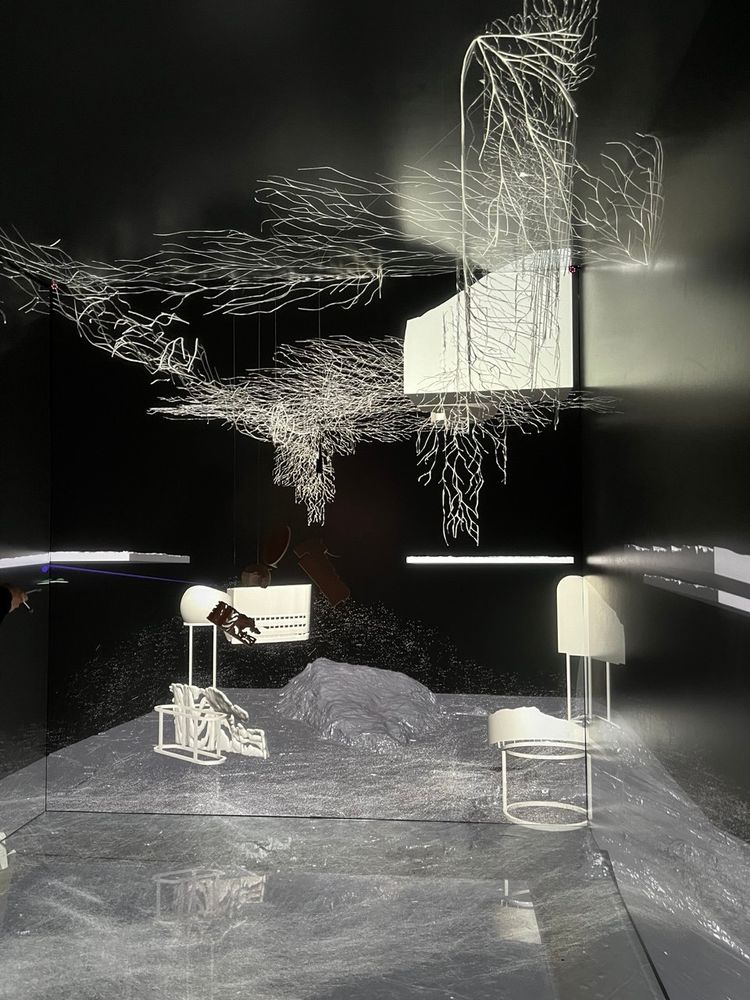 The CAVE with white wireframe-like root structures suspended from the ceiling and abstract, minimalist white sculptures on the floor. The scene is intricately lit, creating ethereal reflections and an otherworldly atmosphere.