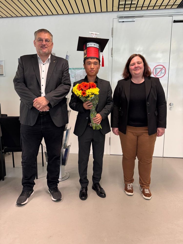 Michael Resch, Qifeng Pan, and Andrea Beck in the entrance hall of HLRS. Qifeng holding flowers and wearing a traditional PhD hat.