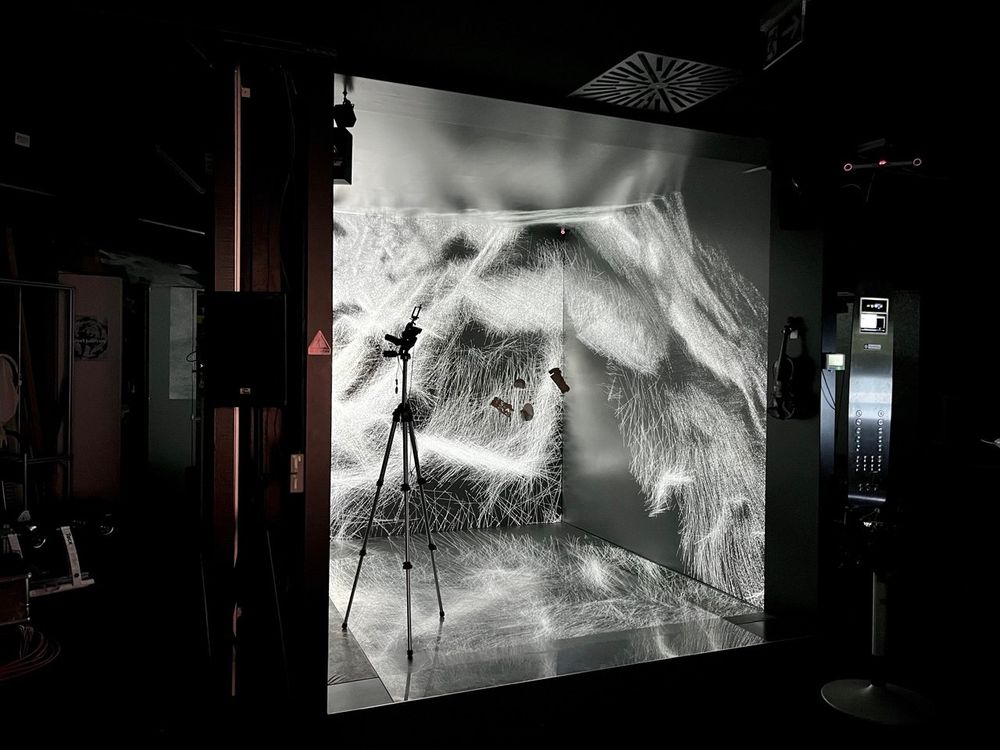 HLRS's CAVE brightly illuminated by white, flowing lines projected onto its walls and floor, creating an immersive, abstract visual resembling a digital meadow or neural network. A tripod with a mounted camera stands inside the space, capturing the projection.