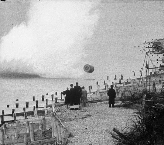 Movie still, showing an inert, practice version of the Upkeep bouncing bomb being dropped during a training flight by members of RAF 617 Squadron at Reculver bombing range, Kent. The bomb's designer, Barnes Wallis, and others watch the practice bomb strike the shoreline. Source: Imperial War Museums, https://commons.wikimedia.org/wiki/File:Bouncing_bomb_training_-_IWM_FLM_2343.jpg