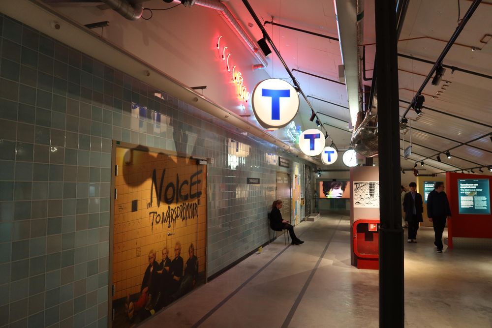 Exhibition with wall covered with turquoise tiles. "T" metro symbol signs hanging from the roof. 