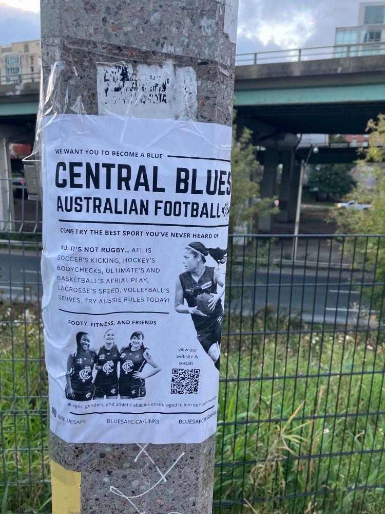 A poster on a lamppost promoting the Central Blues Australian Football Club in Toronto invites people to “try the best sport you’ve never heard of,” describing footy as a mix of soccer’s kicking, hockey’s bodychecks, and volleyball’s serves. It shows three smiling players and a larger image of a player in action holding a football. A highway overpass and city greenery are visible in the background.