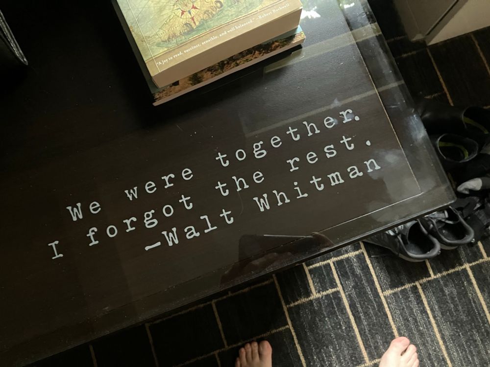 A dark wooden hotel room table with a Walt Whitman quote stenciled in white typewriter font: “We were together. I forgot the rest.” A pair of running shoes sits on the carpeted floor nearby, with a couple of books stacked at the top edge of the table.