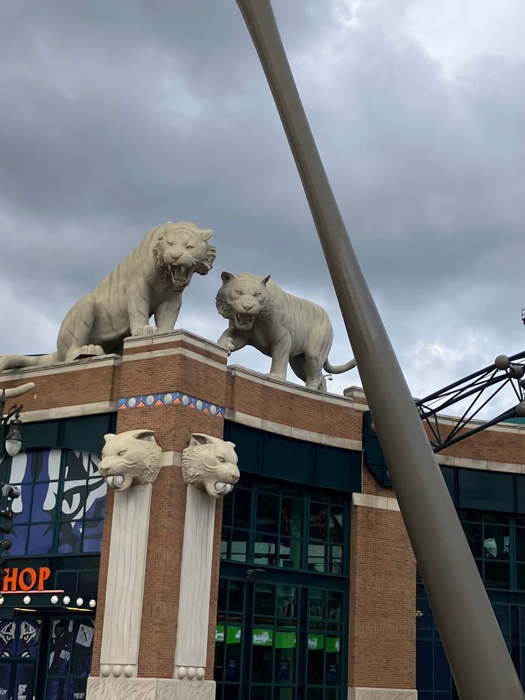 Large stone tiger statues sit on the corner of a brick building under a cloudy sky, appearing to guard the entrance to a stadium in Detroit.