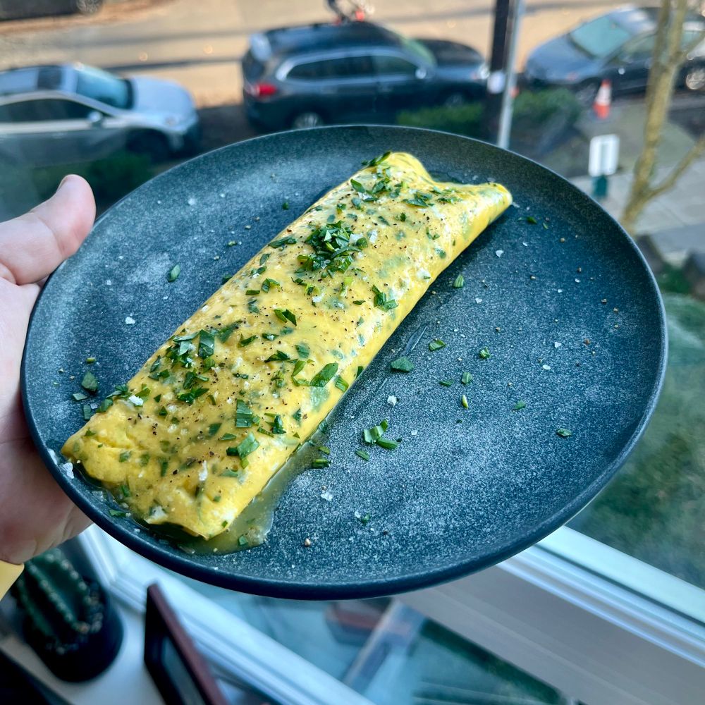 A glossy yellow omelette speckled with green tarragon and some oozy buttery goodness on a grey plate by a bright window. 