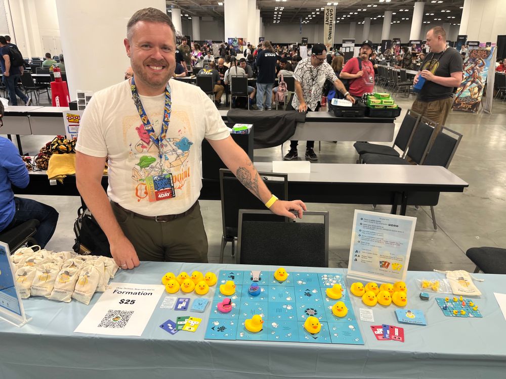 A board game designer with a table full of games and components. A blue table cloth, a number of yellow rubber duckies, cardboard tiles, cards and signs advertising the game Okay, Duckies, Now Let’s Get In Formation. 