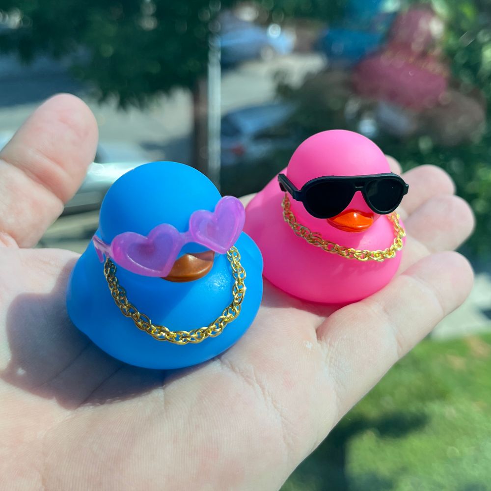 A blue rubber ducky with pink heart sunglasses and a gold chain. A pink ducky with black sunglasses and a gold chain.