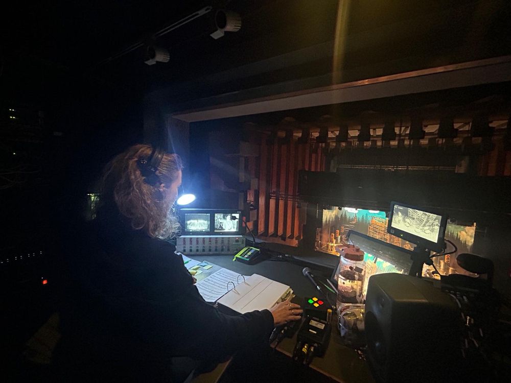 Woman sits at prompt desk looking at prompt book with the cue buttons to her left and auditorium and stage in front of her.