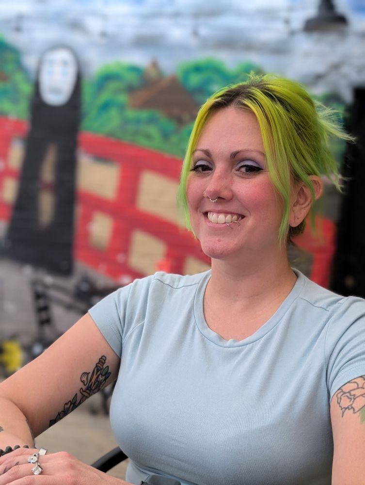 A green haired woman with a mural of No Face from Spirited Away lurking behind her