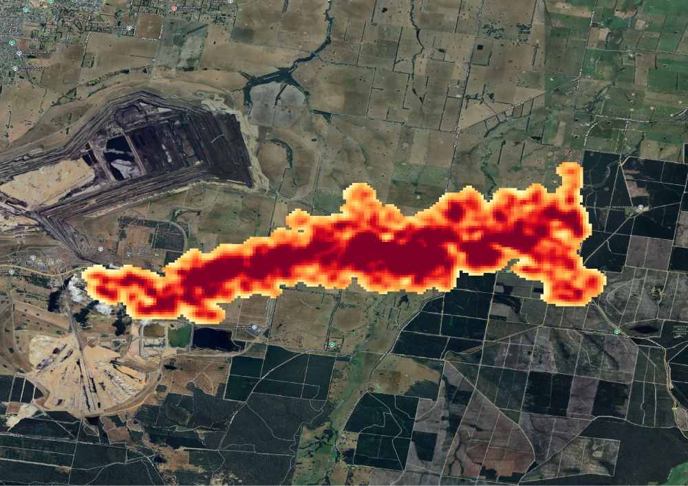 Satellite image of an enormous bright red plume that fades to yellow around the edges. The plume extends ENE from a point on the left hand side of a satellite image of a green landscape. There are two large brown pits on the left of the image.