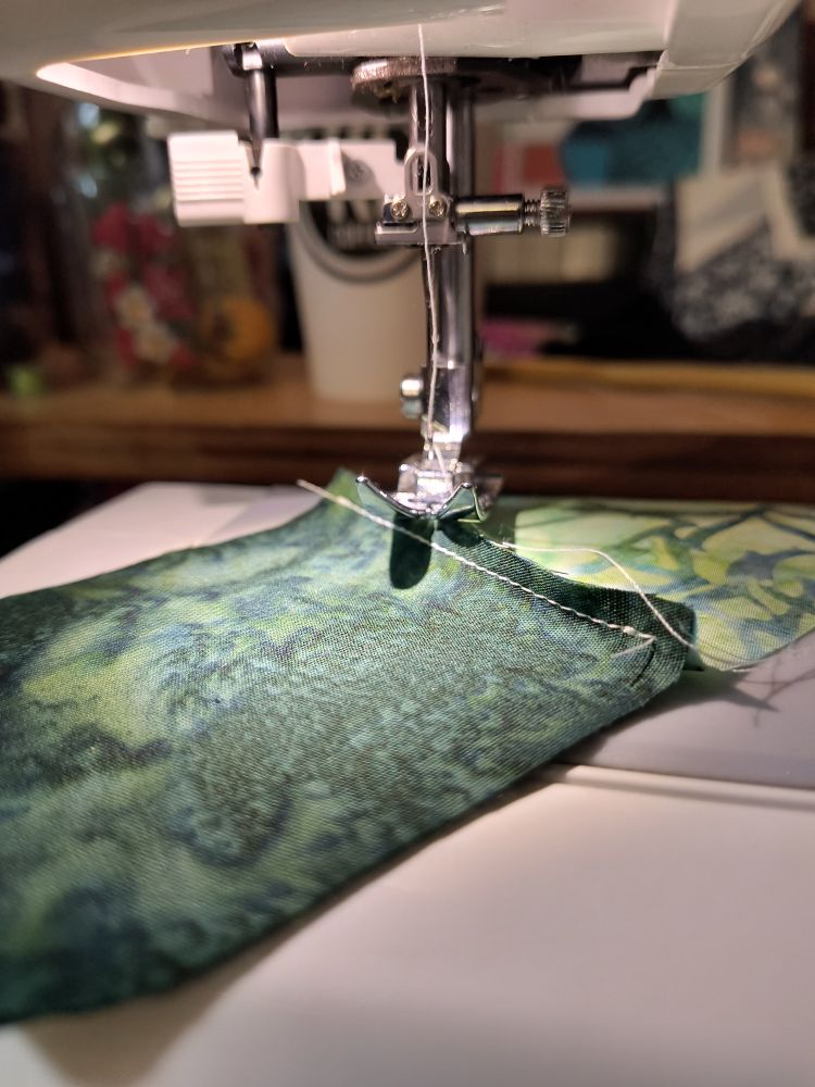 Close up of a sewing machine and fabric. Two squares of different shades of green have been stitched together with white thread.