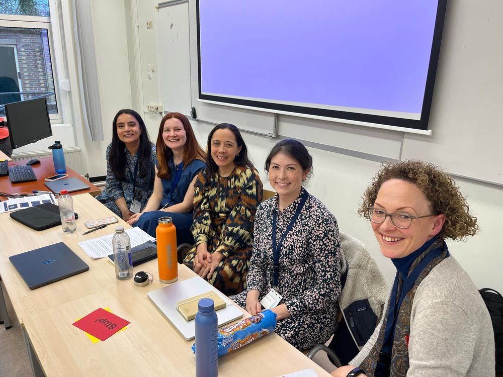 5 women sit smiling in a row behind a desk. A projector screen is on the wall behind them. On the desk in front of them are laptops, notebooks and drink bottles.