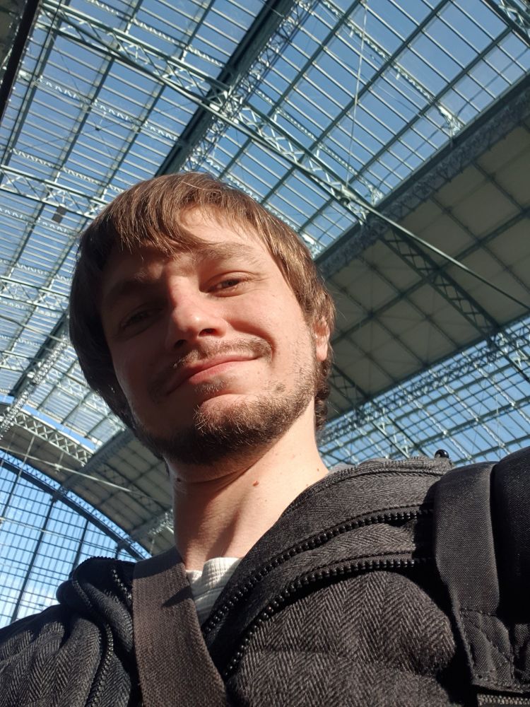 Une photo de ma tête en train de sourire. Visage a moitié éclairé par le soleil derrière on reconnaît la verrière de la gare de Bordeaux. 