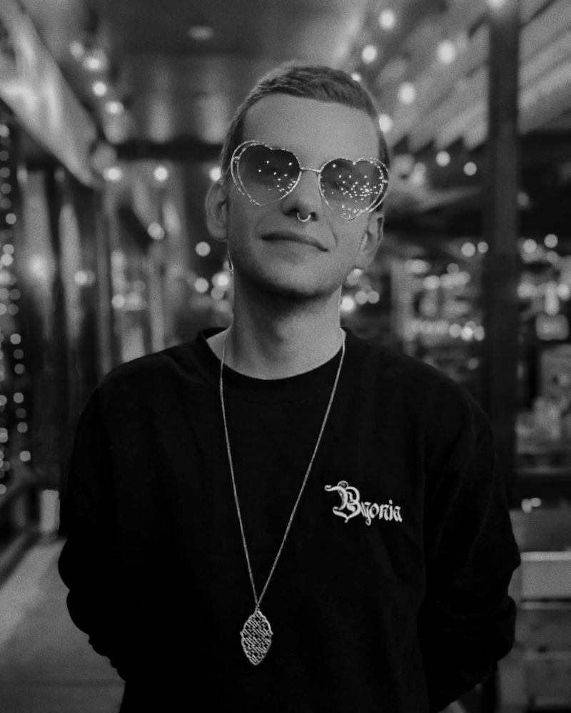 a photo of myself, a male-presenting person in their thirties, with short hair, standing with arms behind my back in a black sweater and a long necklace outside of a well-lit restaurant at nighttime