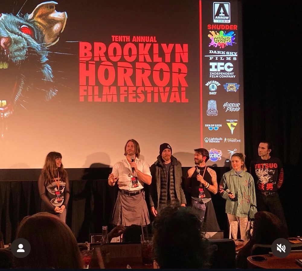 The Tenth Annual Brooklyn Horror Film Festival pre screening intro to the film "It Needs Eyes". Six people stand in front of a film screen with the Brooklyn Horror Film Festival logo, they hold microphones.
