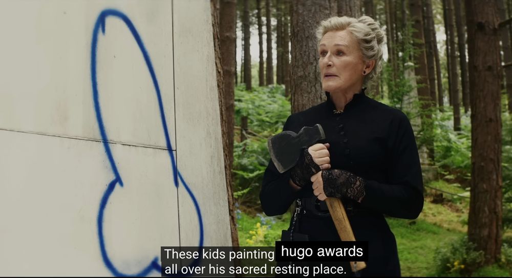 screencap from the trailer of Wake Up Dead Man, featuring Glenn Close holding an axe for personal reasons, standing beside a spray painted phallus on a tomb, with the caption "These kids painting hugo awards all over his sacred resting place"
