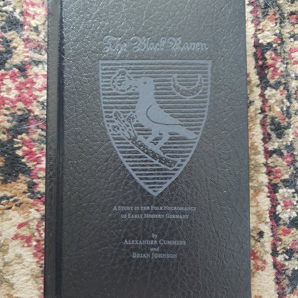 The Black Raven: A Study in the Folk Necromancy of Early Modern Germany by Alexander Cummins and Brian Johnson. A black book on a red and cream carpet. 