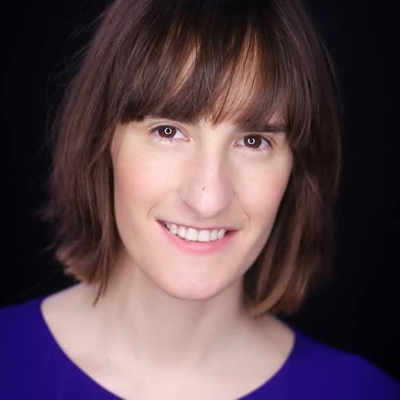 Dr Emma Whipday smiles at the camera against a dark background. She has shoulder-length brown hair and wears a blue jumper.