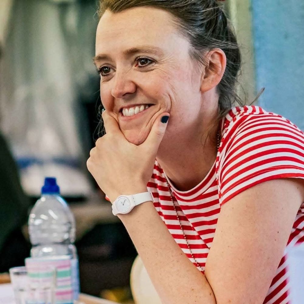Director Amy Leach rests her chin in her hand, grinning at something off camera. She has brown hair and wears a red-and-white striped jumper