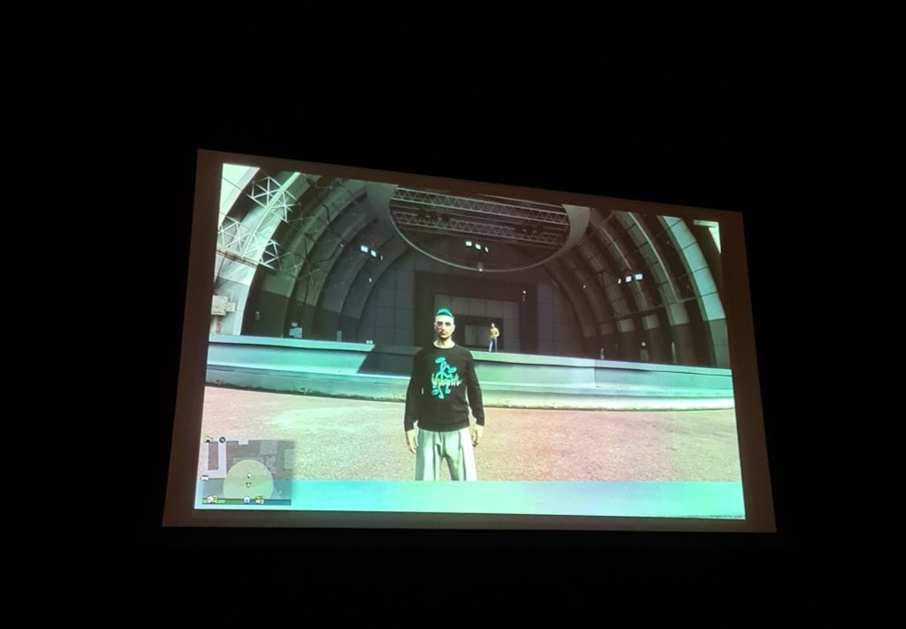 A big screen showing a scene from Grant Theft Hamlet. An actor stands 9n stage in-game in GTA