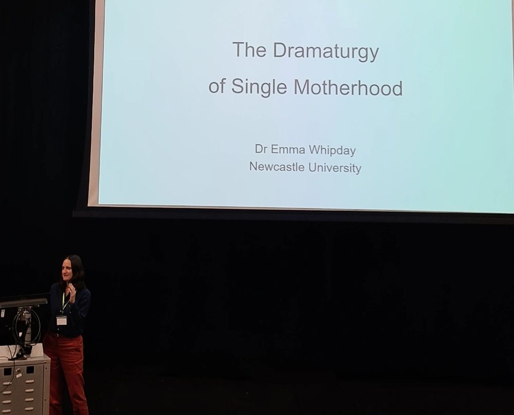 Dr Emma Whipday stands behind a podium before a large screen with the title slide of her presentation on 'the dramaturgy of single motherhood' projected onto it