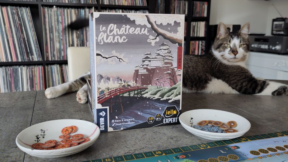 Boite du je Le Château Blanc. Derrière la boîte, couché sur la table, un chat regardant la caméra 