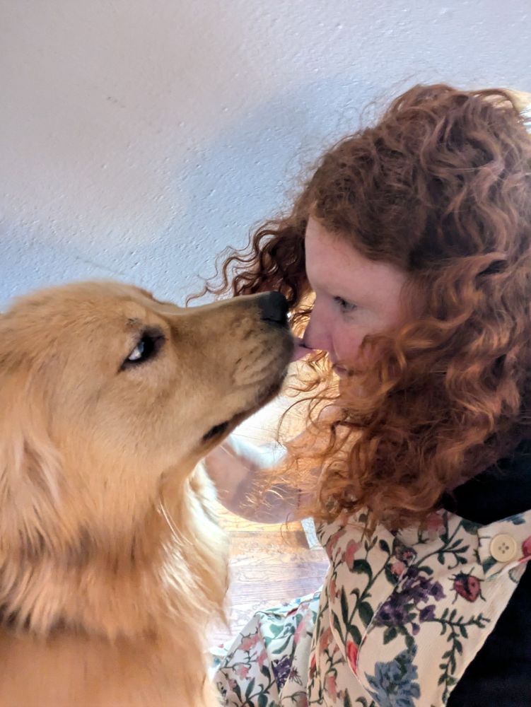 A golden retriever and a reeheaded woman in profile facing each other. The golden retriever has its tongue slightly out and is licking the woman's nose.