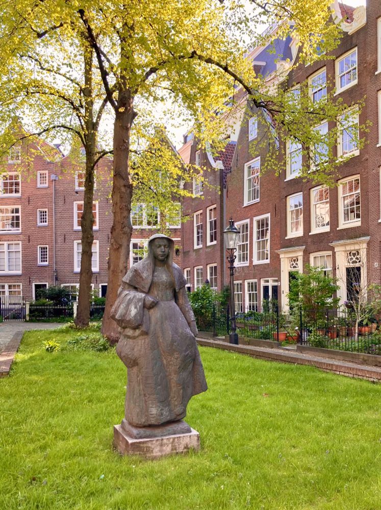 A secluded garden square among old Dutch houses, with a statue of a woman in old-fashioned clothing. There are spring leaves on the trees and the sun is shining.